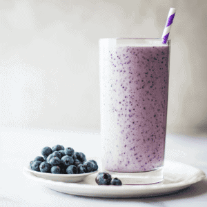 Blueberry smoothie with fresh blueberries on a white plate, served in a tall glass with a purple straw.