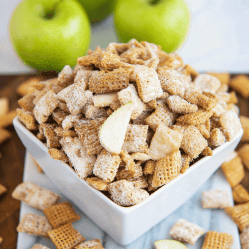 Crunchy apple and graham cracker cereal snack in a white bowl, fresh green apples in background.
