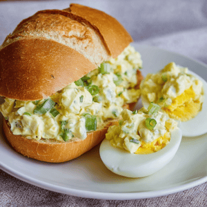 Creamy egg salad sandwich with chopped green onions on a crusty bread roll, served with halved boiled eggs.