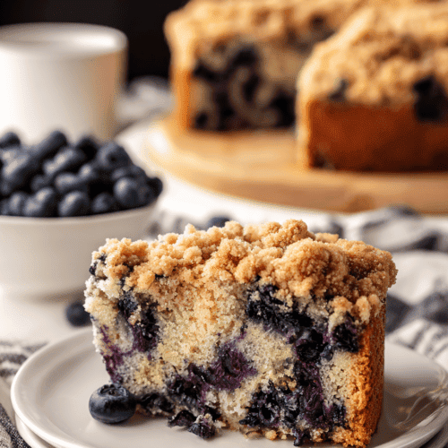 Fresh blueberry crumble cake slice on a white plate with whole blueberries and a whole blueberry crumble cake in the background.