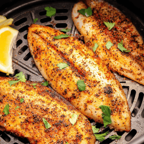 Pan-seared seasoned fish fillets with herbs and lemon wedges in an air fryer.