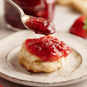 Strawberry jam on a biscuit with a spoonful of jam above, close-up of dessert with fresh strawberries in the background.